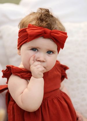 Adorable baby girl with blue eyes wearing a red dress and matching bow headband, with hand near mouth.