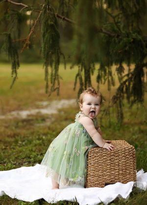 Toddler girl in green tulle dress standing by wicker basket on white blanket under pine tree outdoors.