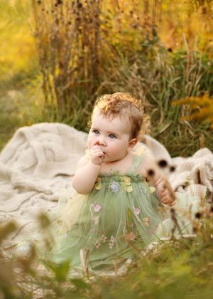 A baby girl in a green floral tulle dress sits on a blanket in a sunlit meadow, hand to mouth.