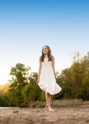 Young girl in white dress walking barefoot on sandy beach with green trees and blue sky background.