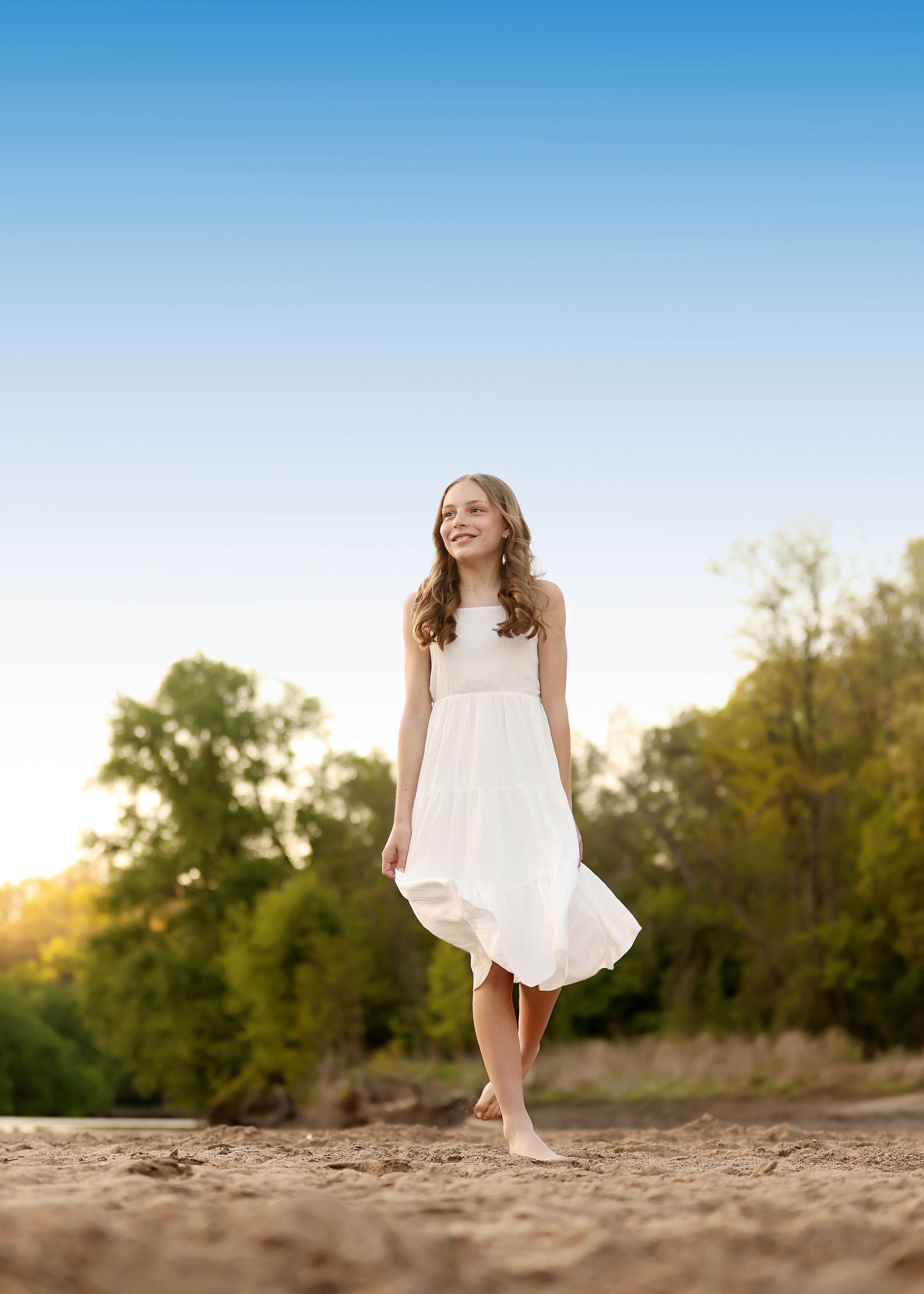Young girl in white dress walking barefoot on sandy beach with green trees and blue sky background.