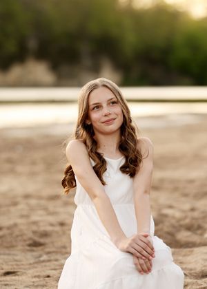 Young girl in white dress sitting on sandy beach at golden hour, smiling with wavy hair.
