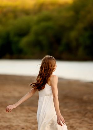 Girl in white dress with long auburn hair walking on sandy beach at golden hour.
