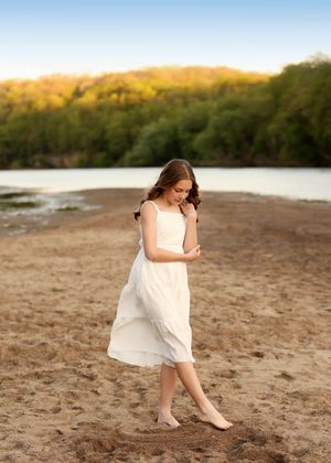 Young girl in white dress walking barefoot on sandy beach with river and green trees in golden hour light.