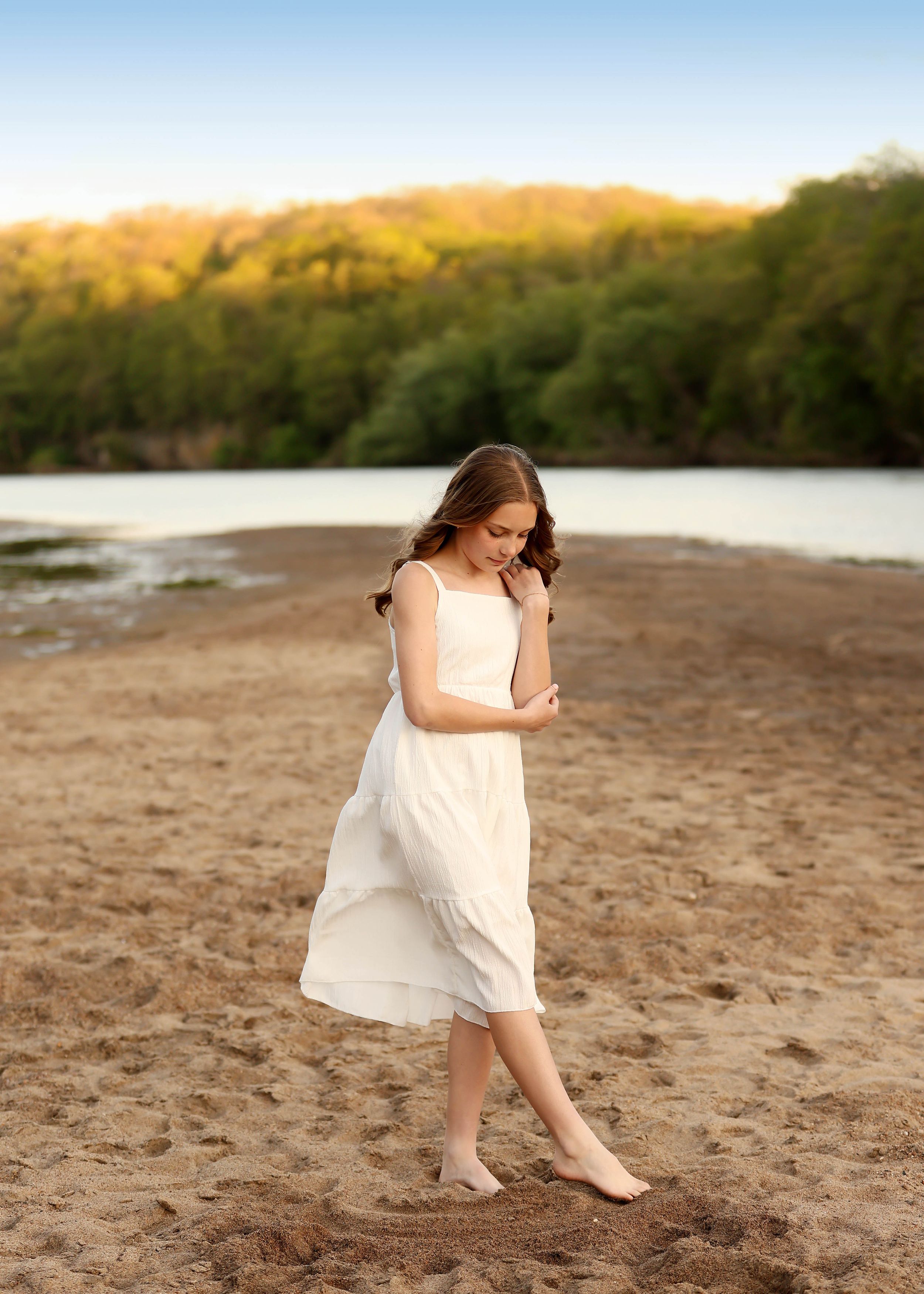Young girl in white dress walking barefoot on sandy beach with river and green trees in golden hour light.