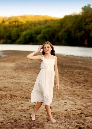 Smiling girl in white sundress standing barefoot on sandy beach at sunset.
