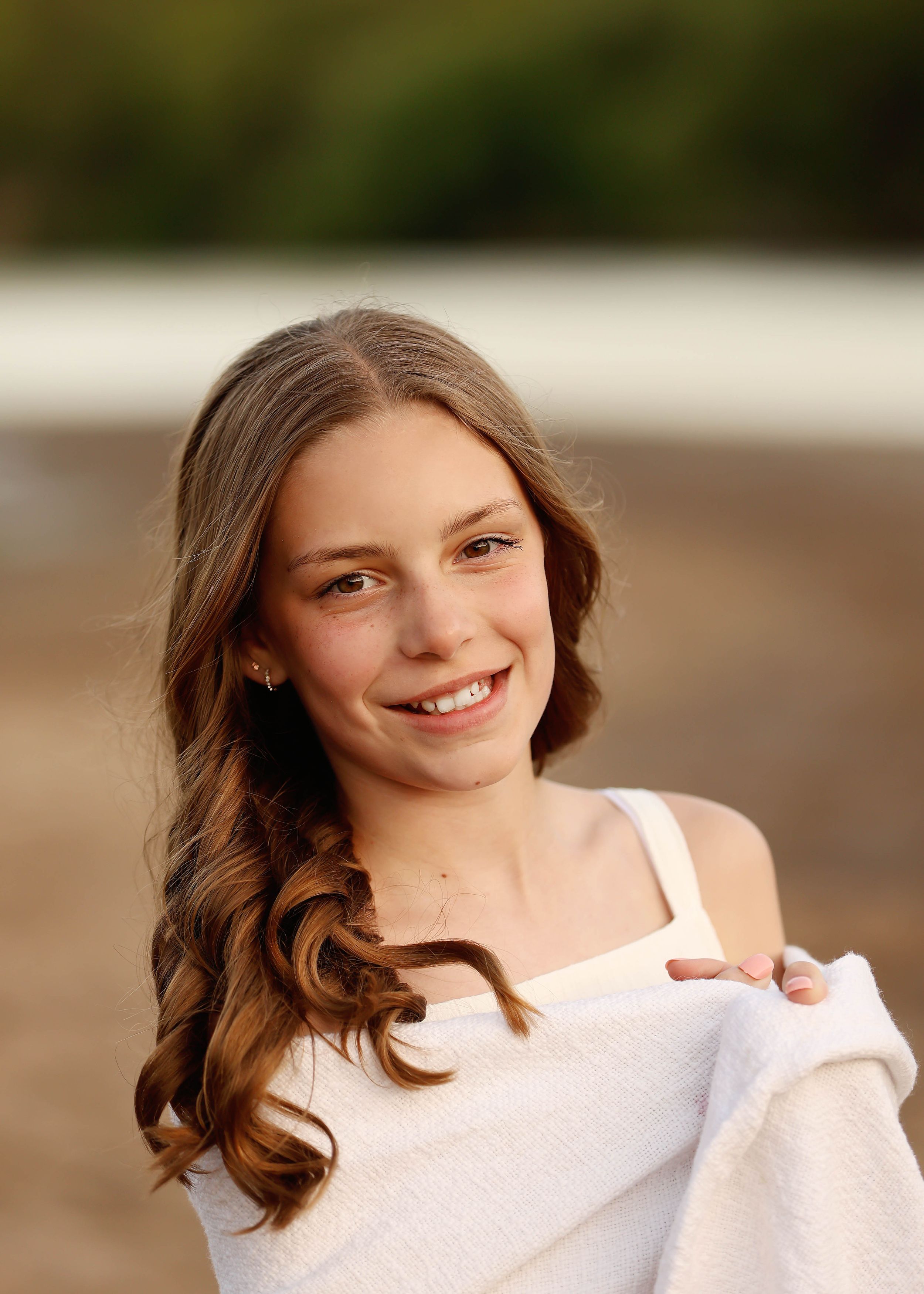 Smiling young woman with long wavy brown hair wearing a white top at a beach.