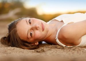 Young woman with brown hair lying on sandy ground, gazing at camera with warm golden sunlight in background.