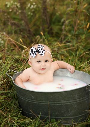A cute baby sits in a rustic galvanized tub filled with milk outdoors, wearing a cow-print bow headband.