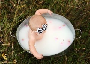 Baby sitting in a galvanized tub filled with milk and pink flower petals outdoors on green grass.