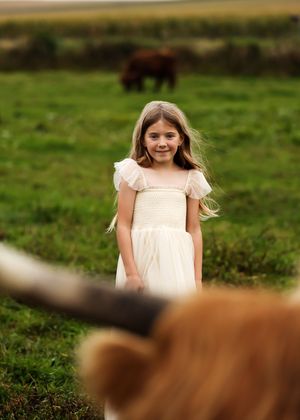 Young girl in white dress smiling in green field with cattle in background and foreground.