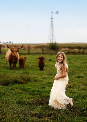 A smiling girl in a white dress runs through a green pasture with Highland cattle and a windmill in the background.