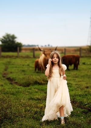 A young girl in a cream dress blows bubbles in a green field with Highland cattle grazing behind her.