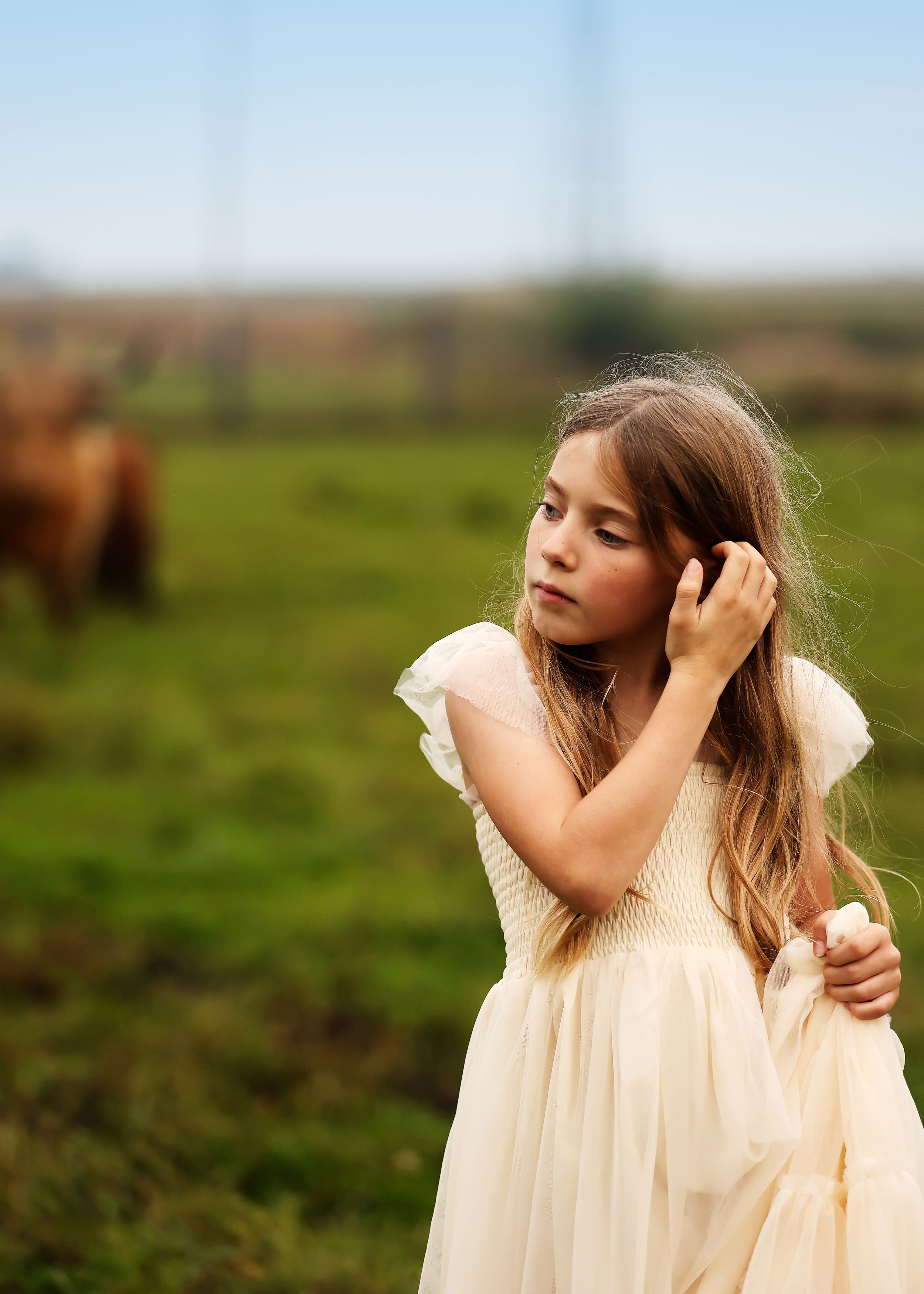 A young girl in a cream tulle dress stands in a green field, gently touching her hair, with a horse in the background.