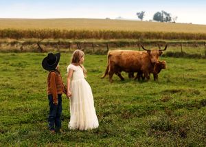 Two children in formal attire hold hands while observing a Highland cow on a lush green farm field.