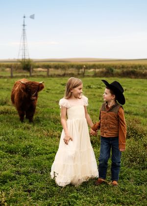 Two children holding hands in a green pasture with a Highland cow and windmill in the background.
