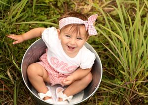 Happy baby girl in pink gingham outfit sitting in metal bucket outdoors, smiling joyfully surrounded by green grass.
