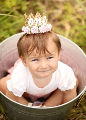 A smiling baby girl in a metal bucket wearing a floral crown that reads one, celebrating her first birthday outdoors.