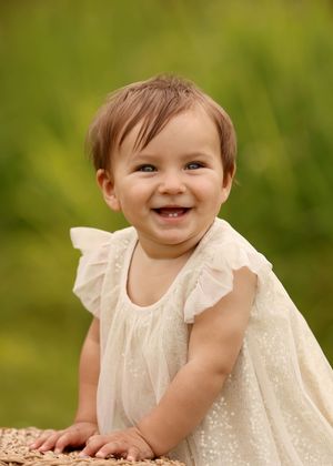 Smiling baby girl in white ruffle dress laughing outdoors with green bokeh background.