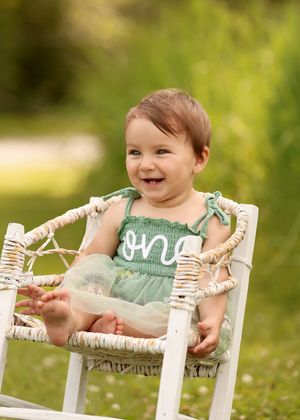 Smiling baby girl in green "one" romper sits in white wicker chair outdoors for first birthday photo session.