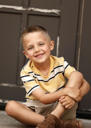 Young boy in yellow striped polo shirt sitting cross-legged on floor, smiling cheerfully at camera.