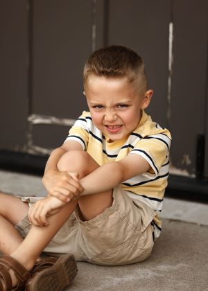 Young boy in yellow striped shirt and khaki shorts sitting on ground, hugging his knee with a grimacing expression.