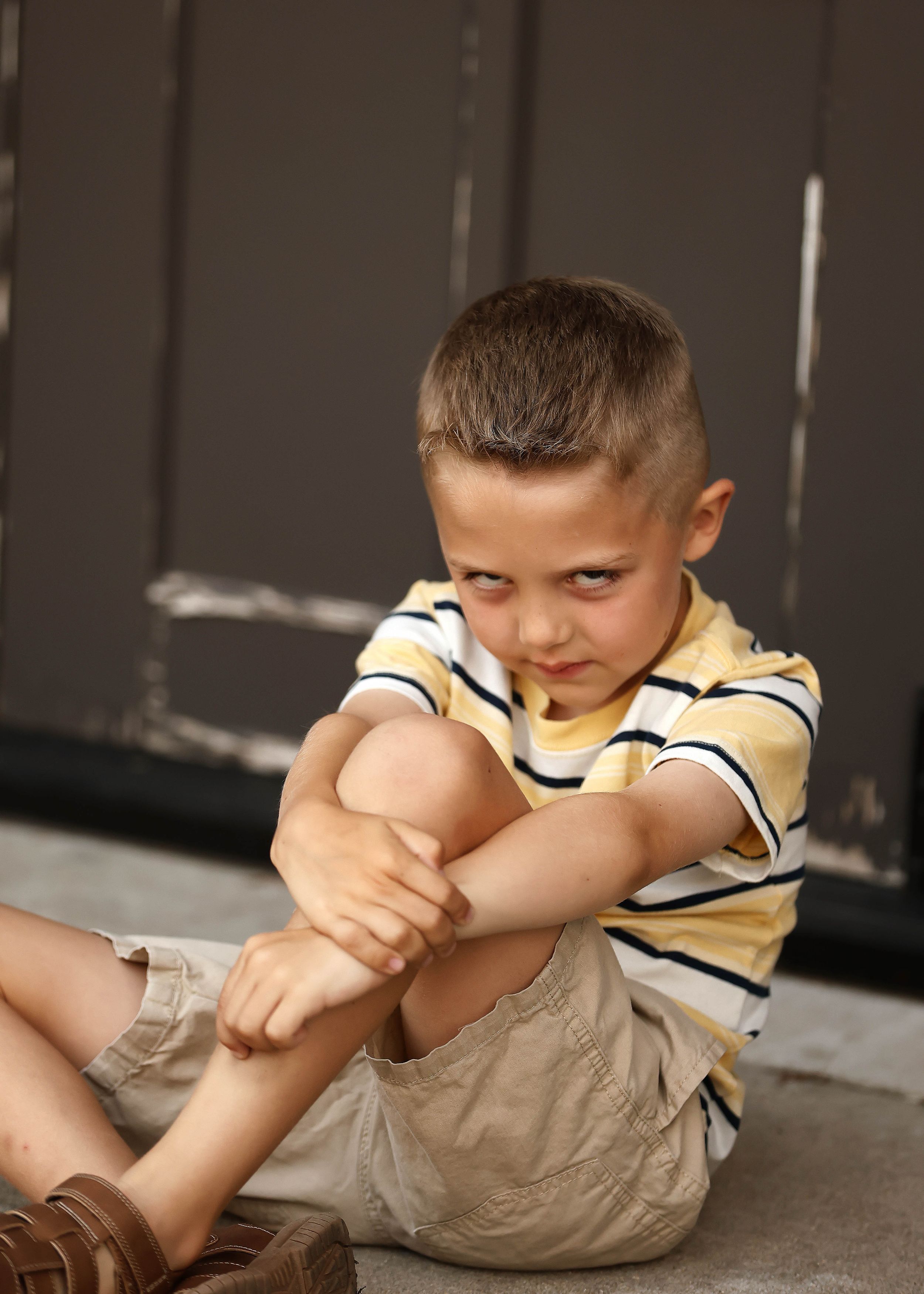 Young boy in striped shirt sitting on ground hugging his knees with a sulking expression.