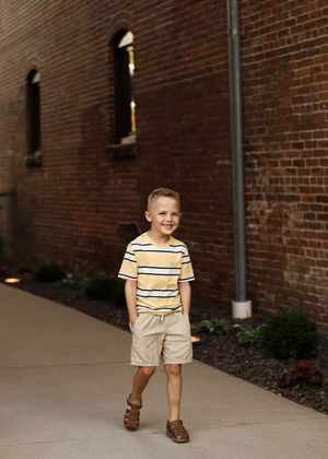 Young boy in yellow striped shirt and khaki shorts smiling outdoors beside a red brick building.