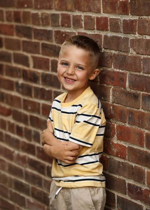 Smiling young boy in yellow striped shirt leaning against a red brick wall with arms crossed.