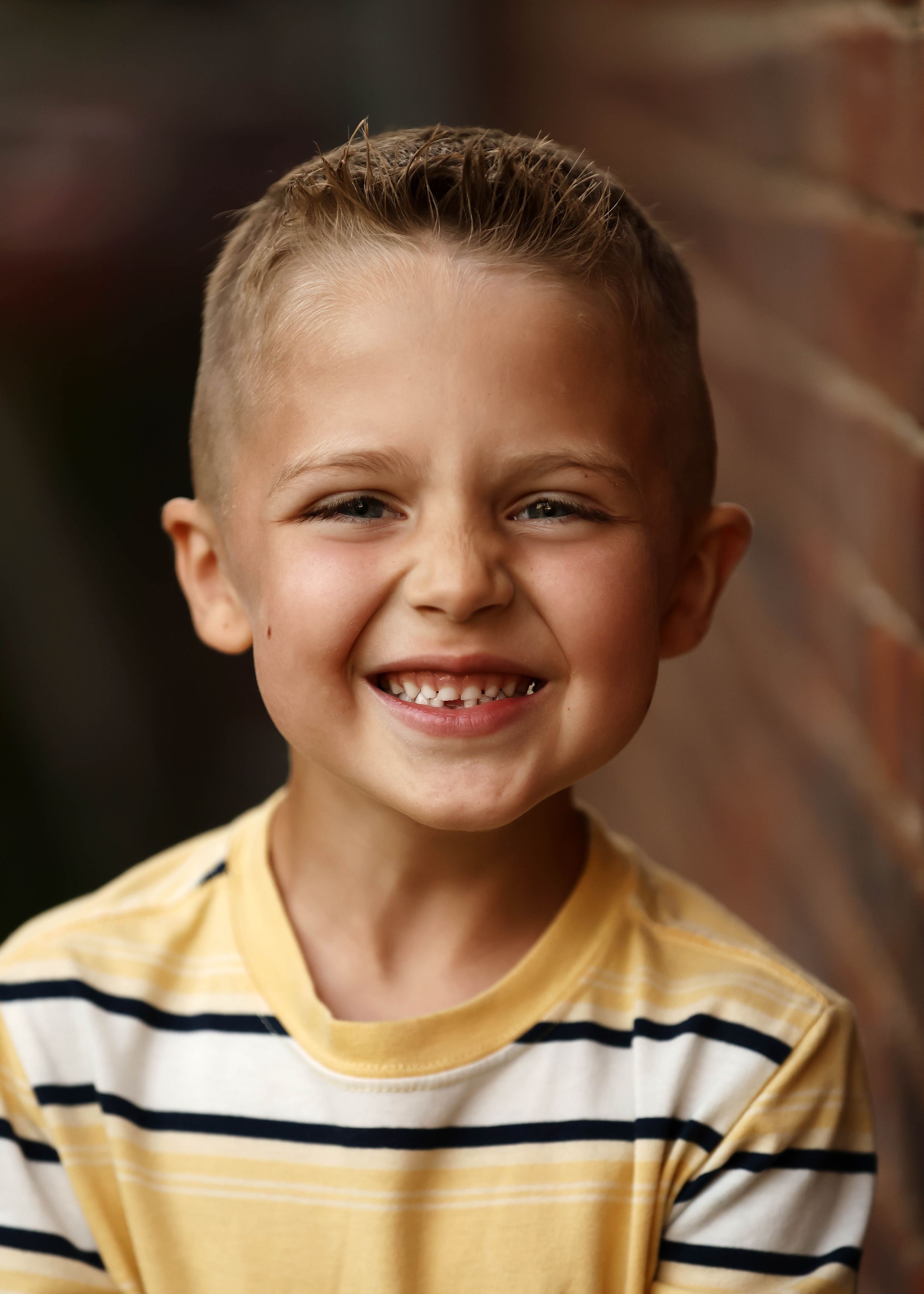 A cheerful young boy with a buzz cut smiles broadly, wearing a yellow striped shirt against a brick wall.