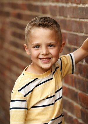 Smiling young boy in yellow striped shirt posing against a brick wall backdrop.