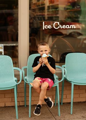 Young boy eating ice cream cone on teal chair outside an ice cream shop.