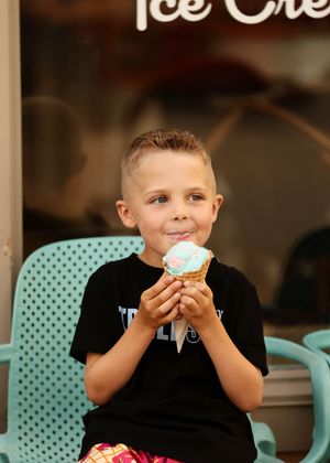 Young boy smiling while holding a colorful ice cream cone in a waffle cone, seated outside an ice cream shop.