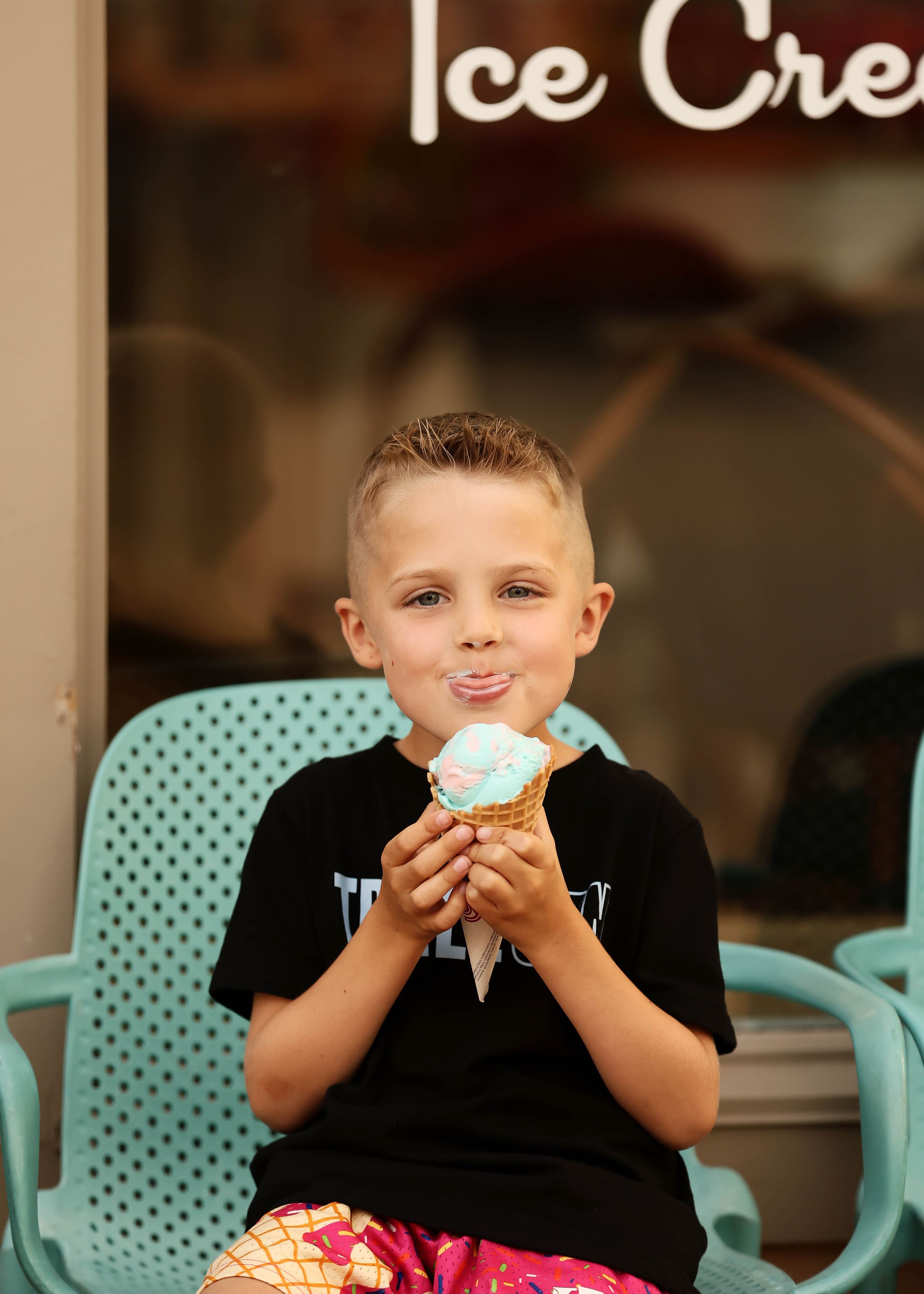 Young boy smiling and enjoying a blue ice cream cone in a waffle cone outside an ice cream shop.