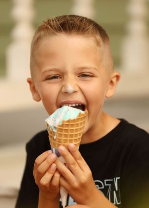 A young boy with a big smile eagerly biting into a blue and pink ice cream cone outdoors.