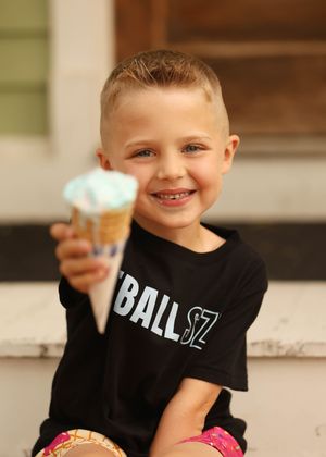 Smiling young boy holding up a blue ice cream cone while wearing a black t-shirt outdoors.