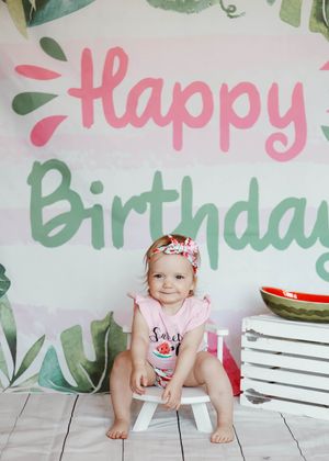 Baby girl in pink outfit sits before a Happy Birthday banner with watermelon decorations at her first birthday party.