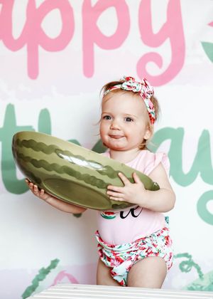 Cute baby girl holding large watermelon bowl at birthday party, wearing floral swimsuit and bow headband.