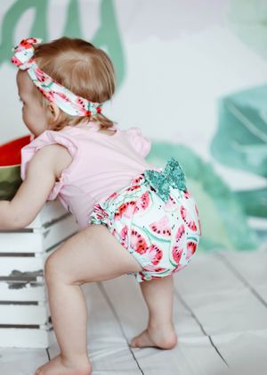 Baby girl in pink top, watermelon print bloomers, and matching bow headband climbing on a white wooden crate.