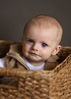 Adorable blue-eyed baby sitting in a woven wicker basket wrapped in a cozy brown blanket.
