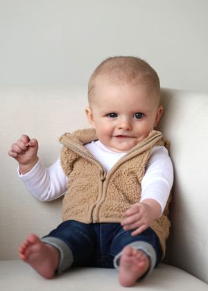 Smiling baby in tan fleece vest and jeans sitting on white chair with one fist raised.