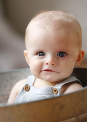 Adorable baby with blue eyes sitting in a metal bucket wearing a light gray knit romper with wooden buttons.