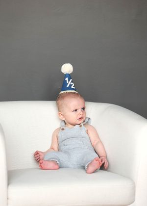 Adorable baby in gray overalls and navy half birthday party hat sits on white chair for 6-month milestone photo.