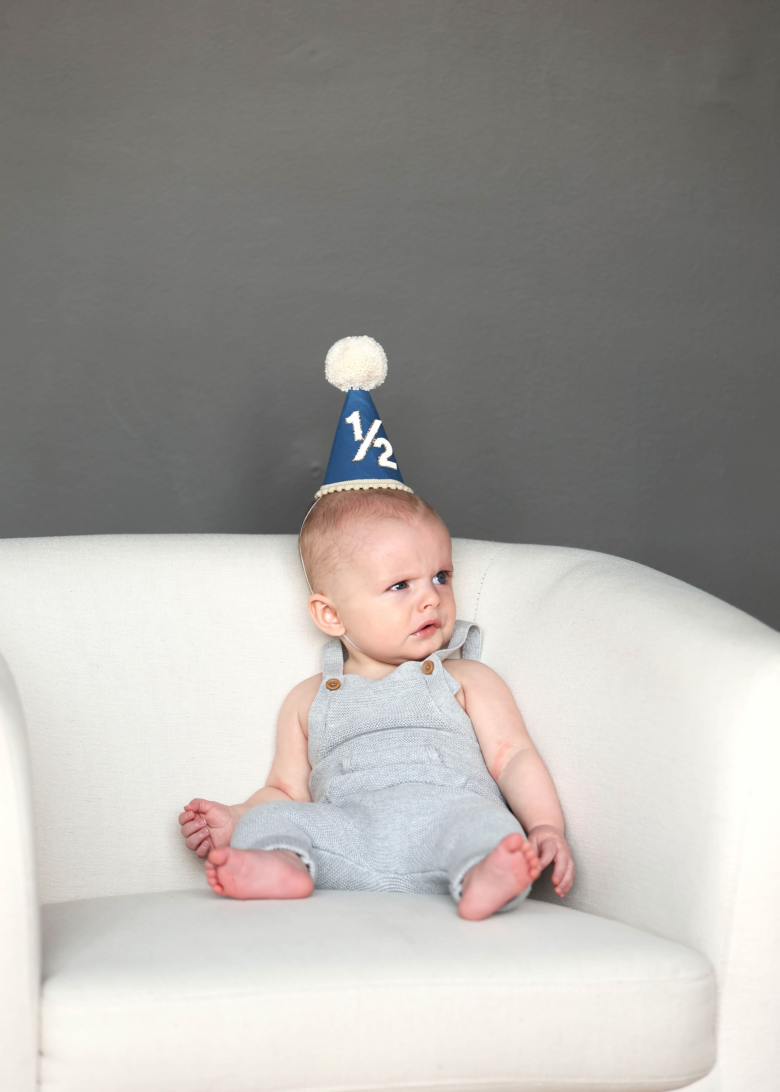 Adorable baby in gray overalls and navy half birthday party hat sits on white chair for 6-month milestone photo.