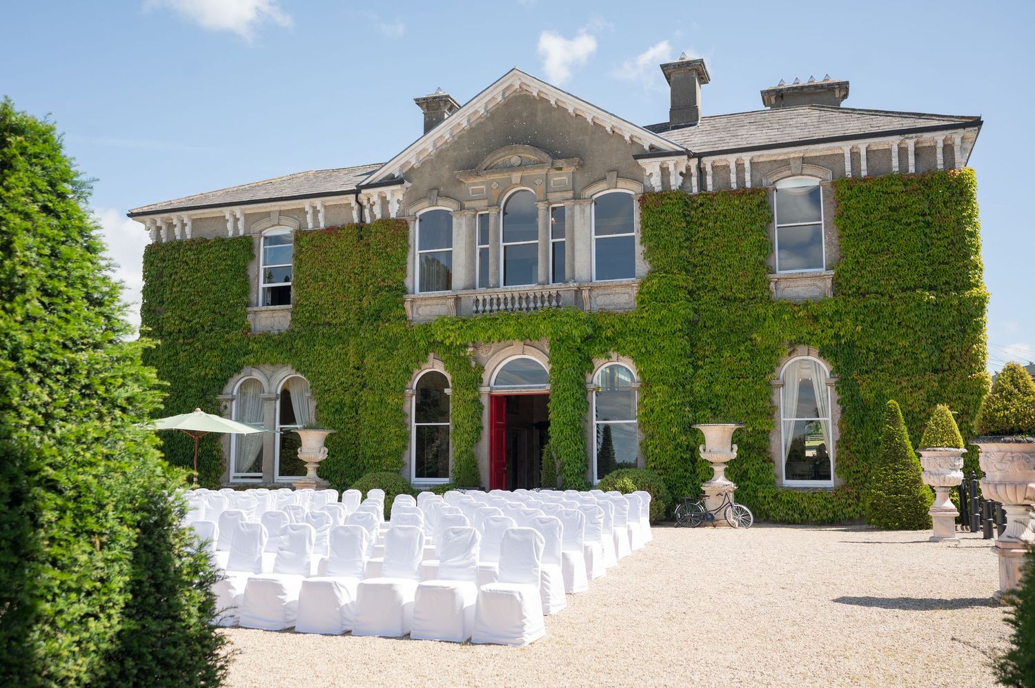 Ivy-covered historic manor house with white chair covers set up for an outdoor wedding ceremony on a sunny day.
