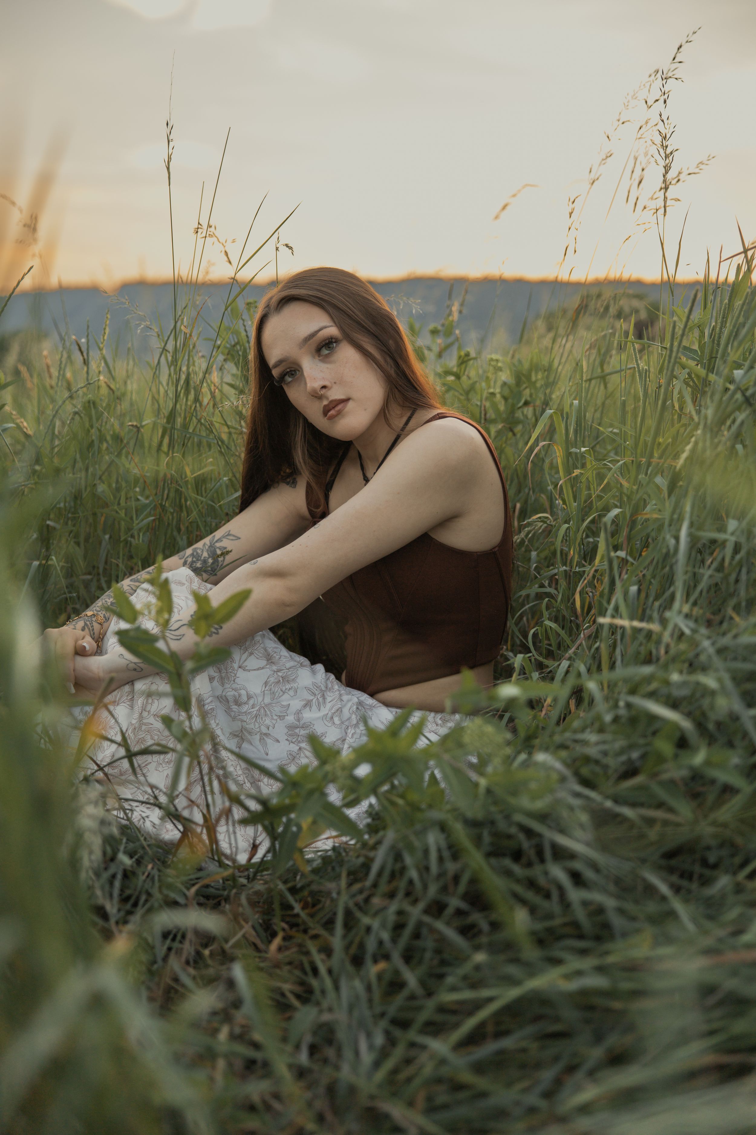 Young woman in brown crop top and white skirt sitting in tall green grass at golden hour.