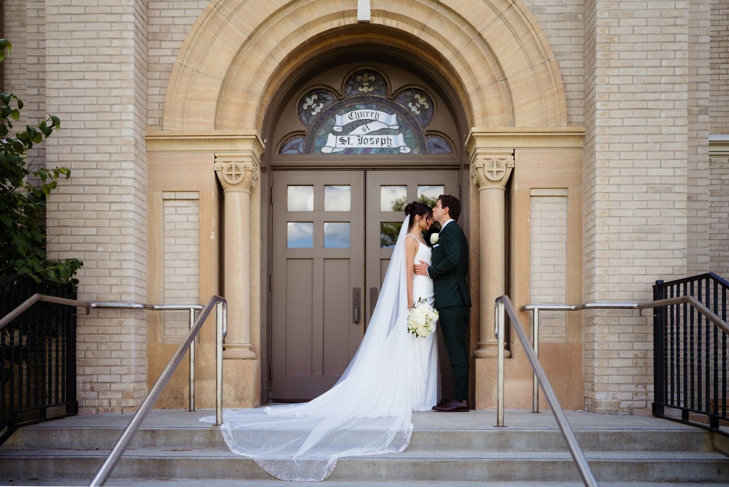 Bride and groom kissing at church entrance with flowing cathedral veil on wedding day.