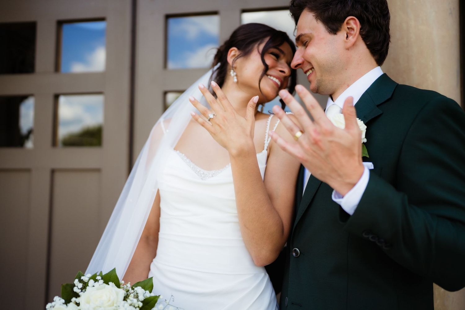 Bride and groom smiling and showing wedding rings, bride holding white bouquet, groom in dark green suit.