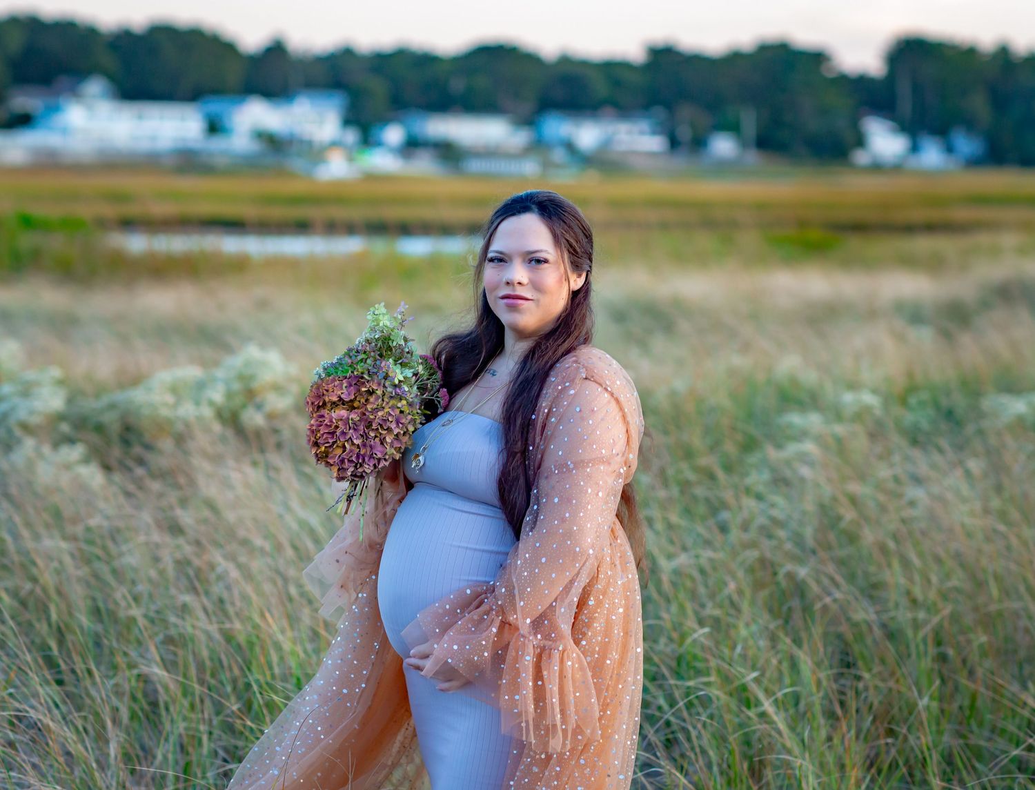 Pregnant woman in gray dress and peach lace robe holding wildflower bouquet in a scenic marsh field at sunset.