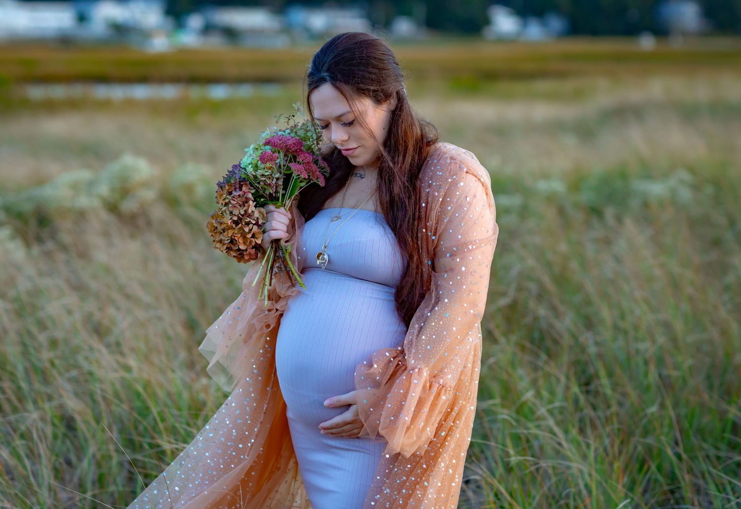 Pregnant woman in blue dress and peach robe holding wildflower bouquet in golden grass field.